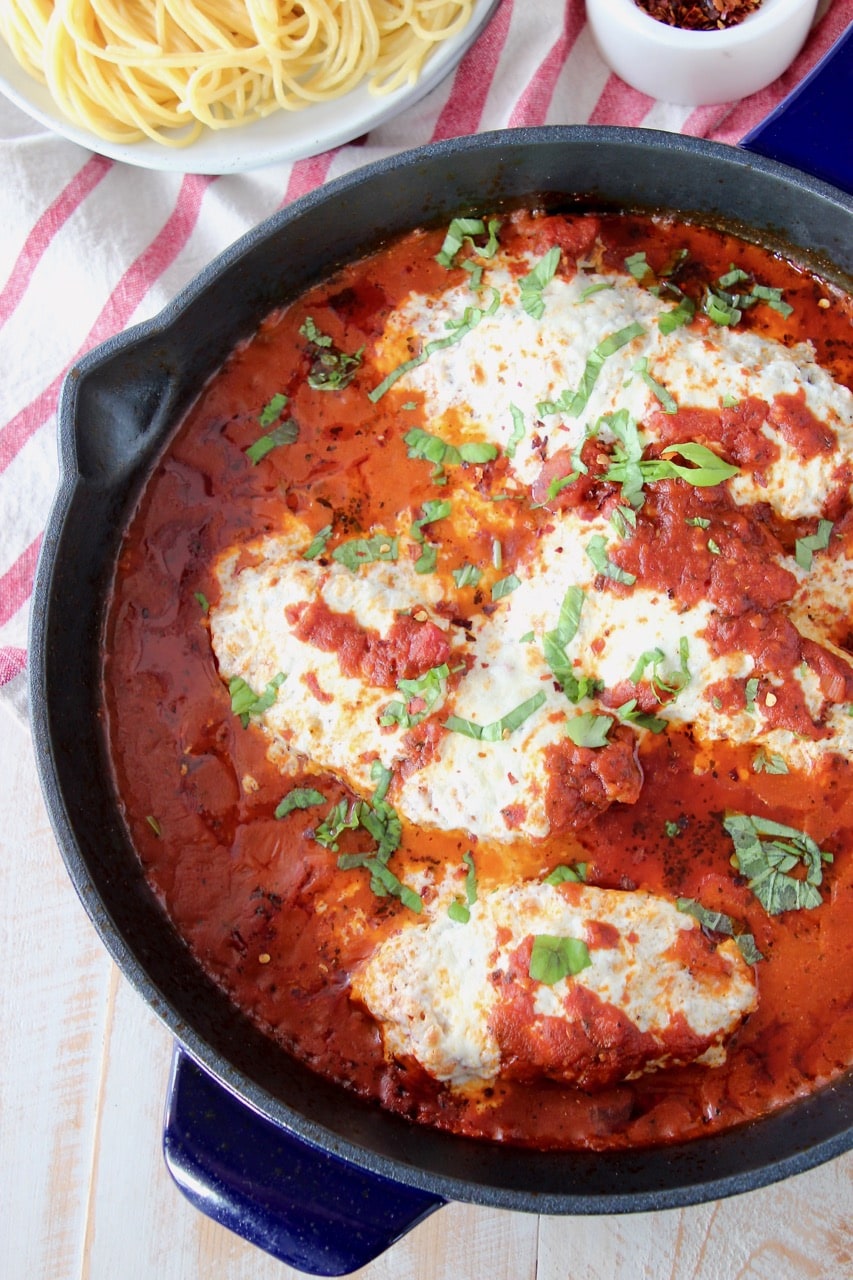 Gluten free chicken parmesan covered in marinara sauce in large cast iron skillet with a bowl of spaghetti noodles on the side