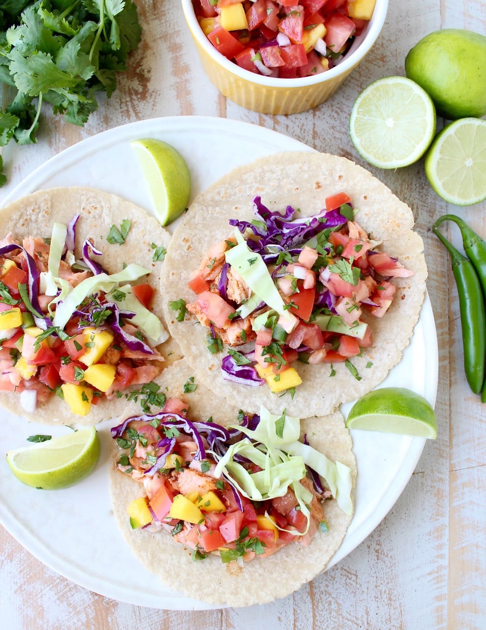 three tacos on a plate topped with salmon, mango salsa and shredded cabbage, with lime wedges on the side