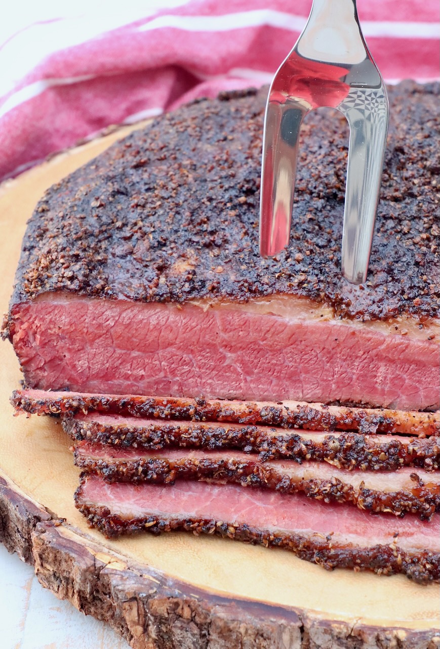 sliced pastrami on wood board with a meat fork stuck in the pastrami