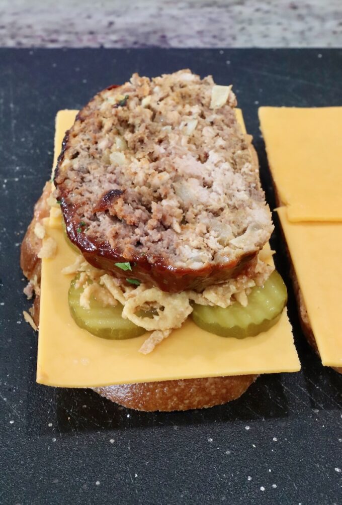 slice of meatloaf on top of cheese, pickles, and fried onion strings, on a piece of bread